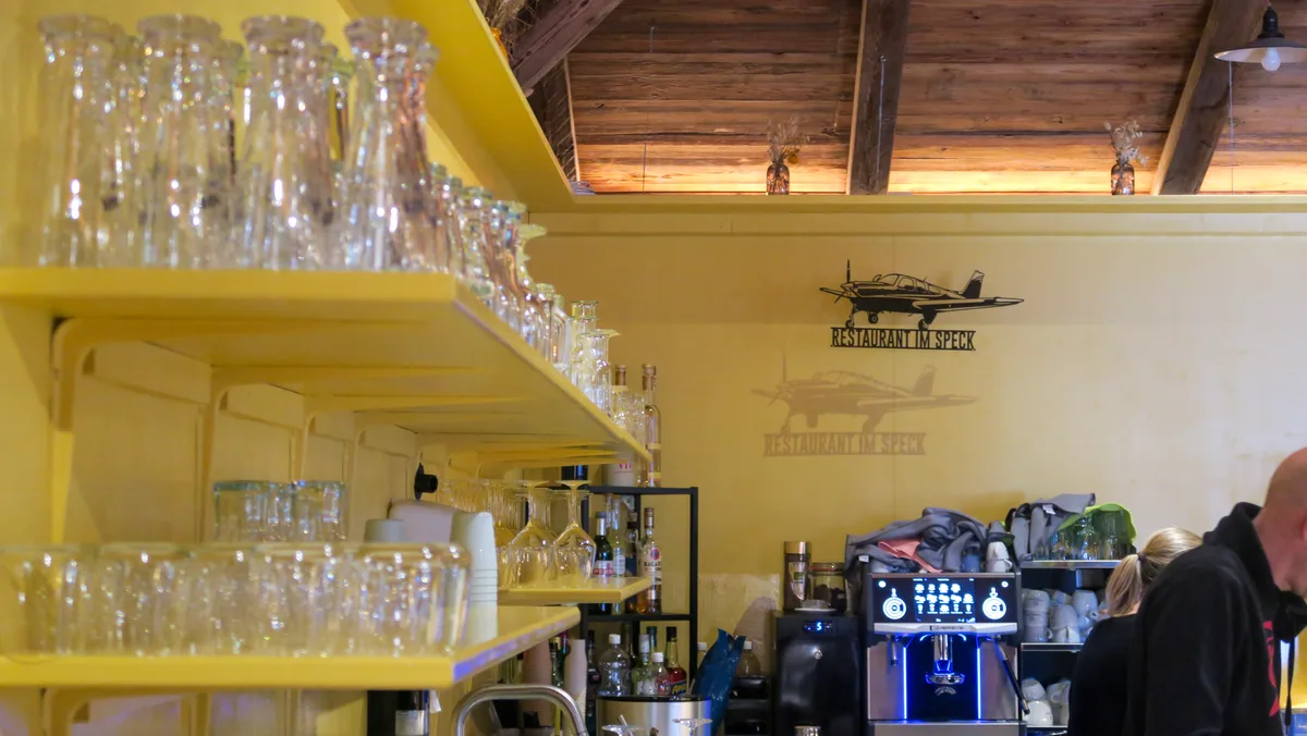 An der Bar des neuen Chalets warten Gläser und Kaffeemaschine darauf, die Wünsche der Gäste zu erfüllen. Man sieht ein Regal, auf dem Gläser stehen, und im Hintergrund eine Kaffeemaschine.